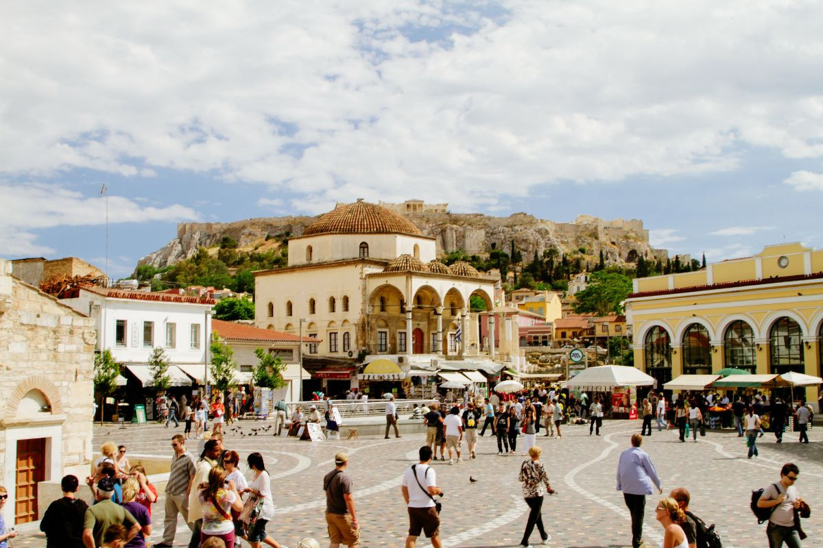 A lively Monastiraki square in Athens with people mingling, vendors under umbrellas, and historical architecture, with a hill in the distance.